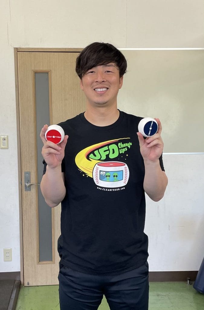 Japanese man smiling with red and blue pokeballs in neutral indoor setting.